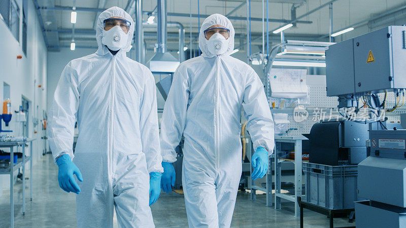 两名工程师/科学家穿着防护服走过技术先进的工厂/实验室。清洁高科技环境与数控机械。